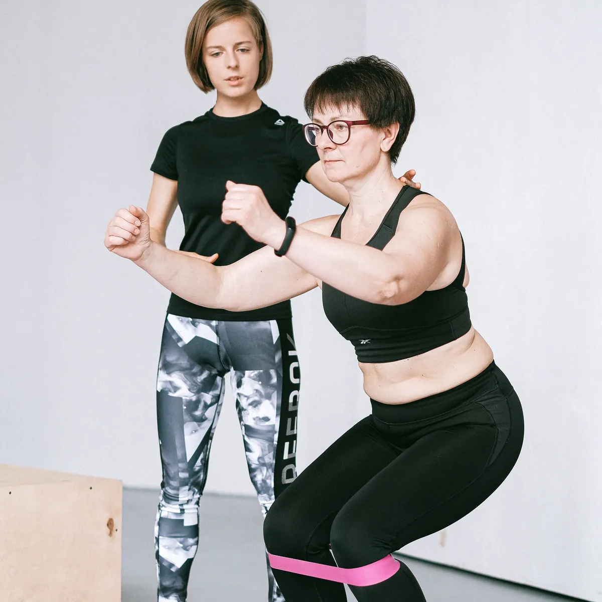 Mujer haciendo sentadillas con banda elástica, acompañada por su entrenadora